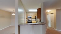 This single-floor residence is designed with an open-concept layout that connects the kitchen to the surrounding living areas, creating a cohesive and functional flow throughout the space. The kitchen serves as a central feature, equipped with stainless steel appliances, including a range, microwave, and refrigerator. Light wood cabinetry lines the walls, providing ample storage, while a taller dark wood cabinet offers a bold contrast. A laminate countertop extends outward to form a raised breakfast bar, which acts as a natural divider between the kitchen and the adjacent living space, making it practical for casual dining or entertaining. The textured ceiling adds a subtle design element consistent throughout the space, and recessed and flush-mounted lighting ensures the kitchen is well-lit. Carpeted flooring extends into the living areas visible from the kitchen, contributing to a warm, comfortable ambiance. Pendant lighting can be seen hanging in the background, adding a decorative touch to the adjacent area. Overall, the space balances practicality with a welcoming, comfortable atmosphere.