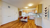 This single-floor residence is designed with a practical and comfortable layout that blends modern finishes with warm, inviting materials throughout. The kitchen serves as a central highlight, featuring light maple cabinetry, granite-style countertops, and a full suite of white appliances including a refrigerator, electric range, microwave, and dishwasher. A freestanding island with seating for two provides an ideal spot for casual dining or meal prep, while warm hardwood flooring ties the kitchen seamlessly into the adjacent living area. The living space is generously sized and bathed in natural light through large vertical-blind windows and a glass door that opens to a private balcony, offering pleasant outdoor views of surrounding greenery. The bedroom is carpeted for comfort and includes a window with outdoor views, built-in storage access, and ample room for furnishings. A second room, also carpeted, offers additional flexible living space. The bathroom is clean and functional, featuring a maple vanity with a white countertop, a large mirror, and a shower-tub combination. Throughout the residence, neutral wall tones, consistent wood accents, and well-placed lighting create a cohesive and welcoming atmosphere.