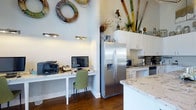 This single-floor residence offers a welcoming and functional layout with a distinct coastal character. The kitchen stands out as a central feature, showcasing crisp white cabinetry, striking marble-style countertops, and stainless steel appliances including a side-by-side refrigerator with water dispenser. A built-in peninsula provides additional prep and gathering space, contributing to the open, airy feel of the area. Adjacent to the kitchen, a dedicated built-in workspace offers a seamlessly integrated home office nook, complete with under-cabinet lighting and ample surface area for a monitor and office equipment — ideal for working from home. Rich hardwood flooring runs throughout, grounding the space with warmth and contrast against the bright white walls and cabinetry. The high ceilings amplify the sense of openness, drawing the eye upward to playful coastal-themed decorative accents, including vintage life rings and colorful oversized lawn game pieces displayed prominently on the upper walls. These distinctive details lend the space a relaxed, resort-inspired personality. Overall, the home balances practical functionality with a cheerful, nautical aesthetic, making it both livable and visually engaging.