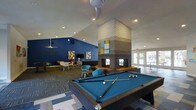 This single-floor recreational space is designed for social engagement and leisure, combining thoughtful design with a lively, energetic atmosphere. The room is anchored by a full-size pool table with a striking blue felt surface, positioned at the center of the space and ready for play. A standout architectural feature is the tall, floor-to-ceiling brick fireplace column that rises dramatically toward the vaulted ceiling, adding a bold structural element that divides and defines the room. Colorful abstract artwork adorns both the fireplace and the deep blue accent walls, contributing to a vibrant, contemporary aesthetic. The lounge area is furnished with mid-century modern-style chairs and small side tables in teal and warm tones, arranged to encourage conversation and relaxation. Recessed lighting throughout the ceiling, complemented by ceiling fans, ensures a well-lit and comfortable environment. A patterned carpet anchors the lounge zone, while a geometric tile floor adds visual interest near the pool table. Large glass doors at the far end of the space invite natural light and provide views of an outdoor area, enhancing the open, welcoming feel of the room.