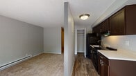 Located at 10306 Meridian Avenue North in Seattle, WA, this single-floor residence offers a straightforward and functional layout throughout. The living area is finished with plush carpeting and neutral gray walls, creating a calm, understated atmosphere. A sliding glass door floods the space with natural light and provides views of the surrounding greenery, enhancing the sense of openness.

The kitchen is efficiently designed with rich dark wood cabinetry, light countertops, and wood-look vinyl flooring that contrasts warmly with the surrounding carpeted areas. Black appliances, including a refrigerator and range, complement the dark cabinetry. A closet adjacent to the kitchen provides convenient additional storage.

The bathroom features dark wood vanity cabinetry with a white countertop sink, a large mirror, wood-look flooring, and a tub-shower combination. Grab bars are installed for added accessibility, and the space is well-lit with wall-mounted fixtures.

Throughout the unit, textured popcorn ceilings and baseboard heating are consistent architectural features. The hallway connecting the rooms is accented with a bold blue-toned wall, adding a touch of visual character. Overall, the space offers a practical and comfortable layout well-suited to everyday living in the heart of Seattle.