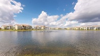 This single-story home is situated within a well-established residential community defined by its picturesque waterfront setting. The standout feature of the surrounding environment is a large, tranquil pond positioned prominently within the community grounds. A decorative fountain at the center of the pond adds visual interest and a sense of movement to the otherwise calm water. The community's architectural style is consistent throughout, with multi-story residential buildings finished in warm, neutral tones and featuring covered balconies and pitched rooflines that reflect a traditional Florida-influenced design aesthetic. Mature landscaping and open green spaces frame the water's edge, contributing to a relaxed, resort-like atmosphere. The expansive sky views and open layout of the community enhance the overall sense of space and airiness. Unfortunately, the interior images provided are too darkly lit to offer meaningful detail about the home's internal layout, finishes, or room configurations. What can be confirmed is that the residence is contained within a single floor, offering the convenience of single-level living within this attractively maintained, water-view community setting.