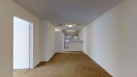 This single-floor space presents an open-concept layout where the living area connects directly to the kitchen, creating a cohesive and functional floor plan. The living area is finished with wall-to-wall carpeting in a warm beige tone, providing a comfortable underfoot feel throughout the main space. Neutral white walls keep the interior bright and versatile, while a textured ceiling adds subtle visual interest overhead.

The kitchen is positioned at the far end of the space and is partially separated by a breakfast bar-style counter, offering a natural division between the cooking and living zones. It is equipped with white appliances, including a range and refrigerator, and features light wood cabinetry that provides ample storage. Under-cabinet lighting illuminates the workspace, while tile flooring in the kitchen area contrasts with the carpeted living space. Ventilation is provided through ceiling registers visible throughout the unit. The overall layout is straightforward and efficient, making good use of the available footprint while maintaining an open, airy feel.