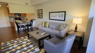 This single-floor space showcases a thoughtfully designed open-concept layout that connects the living area and kitchen into one cohesive environment. The living area is anchored by a gray upholstered sofa accented with green, yellow, and patterned throw pillows, complemented by a matching gray accent chair with white piping detail. A rectangular wood-and-black coffee table sits atop a bold geometric area rug in yellow, gray, and white, adding visual interest to the space. Wall art, including a large horizontal botanical print and a smaller abstract piece, provides decorative character to the neutral-toned walls. A warm table lamp adds ambient lighting to the corner of the room. Adjacent to the living space, the kitchen features a raised breakfast bar with black bar stools, offering a casual dining option. Stone-look countertops and a tile backsplash are visible, contributing to the modern aesthetic. Dark hardwood-style flooring flows throughout, tying the spaces together. The overall ambiance is contemporary and well-coordinated, with a consistent yellow, gray, and green color scheme carried across furnishings and décor.