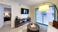 This well-designed single-floor unit showcases a contemporary aesthetic throughout, with light hardwood-style flooring connecting each space cohesively. The open-concept kitchen is a standout feature, equipped with stainless steel appliances including a French door refrigerator, electric range, and built-in microwave. Two-toned cabinetry pairs white high-gloss upper cabinets with rich dark wood lower cabinets, complemented by granite countertops and a neutral tile backsplash. A breakfast bar with pendant lighting provides additional seating and separates the kitchen from the entry corridor, where a sliding-door closet offers convenient storage.

The living area is bathed in natural light through a large sliding glass door that opens to an outdoor space, with light-toned walls and hardwood-style flooring enhancing the airy feel. Both bedrooms feature windows with views of the surrounding building exterior and are finished with the same consistent flooring. One bedroom is styled with a neutral accent wall and warm ambient lighting, while the other maintains a clean, bright palette.

The bathroom is elegantly appointed with large-format white floor tiles, a walk-in tiled shower, and a floating vanity with dual sinks beneath a wide illuminated mirror, creating a calm, spa-inspired atmosphere.