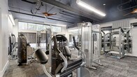 Located at 425 N Boylan Avenue in Raleigh, NC, this expansive fitness center spans a single floor and offers a comprehensive workout environment designed to accommodate a wide variety of training styles. The space features an industrial-modern aesthetic, characterized by exposed ductwork and piping along dark-painted ceilings, elegant wood-bladed ceiling fans, and bold linear pendant lighting that illuminates the floor throughout.

The main workout area is generously proportioned, housing an extensive selection of selectorized strength machines, cable systems, and free weights including a full rack of dumbbells and colorful kettlebells. A dedicated cardio zone features spin bikes, stair climbers, rowing machines, and ellipticals. One section of the fitness center includes a mirrored studio space equipped with resistance bands, stability balls, and additional accessories, complemented by built-in cabinetry and a mounted display screen — ideal for group training or personal sessions.

Patterned carpet tiles in grey and teal tones cover the floors throughout, adding a modern touch while providing comfort underfoot. Large windows along the perimeter invite natural light and offer views of the surrounding neighborhood. A dramatic feature wall adorned with athletic-themed artwork anchors one end of the strength training area, contributing to the space's motivating and polished ambiance.