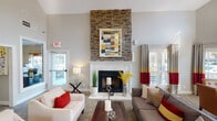 This well-appointed single-floor community space offers a welcoming and sophisticated atmosphere from the moment you enter. The centerpiece of the main lounge is a striking floor-to-ceiling stacked-stone fireplace, crowned with a white marble surround and mantle, and accented by a bold geometric art piece. Soaring vaulted ceilings amplify the sense of openness, while large windows fitted with white plantation shutters and color-blocked curtains in red, yellow, and gray flood the interior with natural light and frame views of the surrounding greenery. The main seating area is furnished with plush sofas and accent chairs arranged around a rich wood coffee table, creating an inviting space for gathering. An adjacent lounge area features additional seating, a glass-top dining table with leather chairs, and continued vaulted ceilings that enhance the airy feel. A private office, visible through an interior glass window, provides a professional workspace tucked seamlessly into the layout. Throughout the space, consistent warm-toned carpeting, neutral gray walls, and carefully curated décor—including decorative lamps, plants, and art—tie each zone together into a cohesive, modern aesthetic.