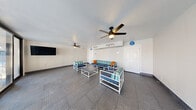 Located at 2301 S Valley View Blvd in Las Vegas, NV, this poolside lounge serves as a comfortable communal gathering space with a welcoming, resort-inspired atmosphere. The single-floor layout makes efficient use of its footprint, offering an open, unobstructed interior that encourages relaxed socializing. The space features textured diamond-plate flooring throughout, lending a clean and durable finish well-suited to a poolside environment. A seating arrangement of white-framed furniture with navy and teal striped cushions, accented by bright orange throw pillows, anchors the room with a fresh, coastal aesthetic. A low-profile coffee table sits at the center of the grouping, complementing the casual lounge setting. Two ceiling fans with integrated light fixtures ensure comfortable airflow throughout the space, an essential feature given the Las Vegas climate. A wall-mounted flat-screen television adds an entertainment element, making the lounge ideal for group gatherings. The interior benefits from abundant natural light, courtesy of large black-framed sliding glass doors spanning one wall, which open directly to the pool deck and reveal views of the outdoor pool area with its arched architectural details. Restroom access is conveniently available within the space, and climate control is provided via wall-mounted HVAC units. Overall, the lounge offers a functional and stylish retreat between the indoors and the pool area.