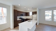 This single-floor space showcases a thoughtfully designed kitchen that serves as the heart of the layout. The kitchen features rich, dark espresso-toned cabinetry that lines the walls in an L-shaped configuration, providing generous storage and a sleek, contemporary aesthetic. The cabinetry is complemented by clean white countertops that extend throughout, creating a striking contrast.

A central island anchors the space, finished in bright white quartz with a double undermount sink, offering additional workspace and a natural gathering point. Stainless steel appliances, including a gas range, over-the-range microwave, and a full-size refrigerator, add a professional quality to the kitchen.

The flooring consists of light-toned wood-look planks that run throughout, contributing warmth and continuity to the space. Recessed lighting in the white ceiling ensures even, practical illumination, while a window above the counter allows natural light to filter in. The overall design is clean and modern, balancing functional efficiency with a polished, contemporary finish.