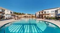 Located at Park Commons in Knightdale, NC, this outdoor amenity area is centered around a striking dual-pool design, where two generously sized pools flow together to create an expansive, resort-style aquatic experience. The crystal-clear water is complemented by a wide, well-maintained concrete deck that provides ample space for lounging and socializing.

Multiple wooden pergola structures are positioned throughout the area, offering shaded retreats furnished with comfortable chaise lounge chairs. Stone-accented columns add a refined architectural touch to the pergolas, blending natural textures with the clean lines of the surrounding hardscape. Additional seating options, including chairs with vibrant red cushions, are thoughtfully arranged to accommodate gatherings of various sizes.

The pool area is enclosed by sleek wrought iron fencing, ensuring both security and unobstructed views of the surrounding multi-story residential buildings. Mature trees and landscaped greenery frame the perimeter, providing a natural backdrop under the open sky. The space is well-suited for both leisurely relaxation and active outdoor enjoyment, offering a central community gathering point. The overall design balances modern amenities with warm, inviting materials, creating a welcoming outdoor environment for residents of the Park Commons community.