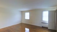 This single-floor space presents a straightforward and functional layout highlighted by rich, polished hardwood floors that run throughout the main living area. The floors exhibit a warm, deep tone and a reflective sheen that adds a sense of warmth and character to the room. Walls are finished in a soft, neutral off-white, providing a bright and clean backdrop that complements the natural materials underfoot. Two windows allow natural light to filter into the space, keeping the interior feeling open and airy. A baseboard radiator is positioned beneath one of the windows, indicating a traditional heating system. The ceiling is smooth and painted white, maintaining a simple, uncluttered aesthetic throughout. White baseboards frame the perimeter of the room, adding a crisp finishing detail that ties the space together. The overall ambiance is modest and understated, well-suited for a variety of uses. The absence of built-in features keeps the space highly adaptable, allowing for flexible furniture arrangements to suit individual needs.
