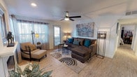 This single-floor space offers a comfortable and thoughtfully designed living area that combines modern style with a warm, inviting atmosphere. The living room serves as the heart of the home, showcasing wood-look plank flooring that adds warmth and a clean aesthetic throughout. A ceiling fan with an integrated light fixture is centrally positioned, providing both airflow and overhead illumination. Natural light filters in generously through multiple windows dressed with sheer curtains and horizontal blinds, creating a bright and airy feel. The living area is furnished with a large, plush sectional sofa in a rich charcoal tone, accented with teal and patterned throw pillows that add a pop of color. A patterned area rug with circular motifs anchors the seating arrangement, complementing the neutral tones of the space. Additional seating is provided by an accent chair with an ottoman, positioned near a corner floor lamp. A bold, abstract canvas painting adorns the main wall, serving as a striking focal point. Greenery from indoor plants adds a natural, refreshing touch to the overall décor. The space also features a door providing direct access to the exterior, enhancing connectivity between indoor and outdoor areas.
