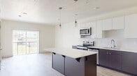 This well-designed 1-bedroom, 1-bathroom unit offers a thoughtful, efficient layout across a single floor. The open-concept kitchen serves as the heart of the space, showcasing a two-tone design with crisp white upper cabinetry and dark lower cabinets. A central island with a white quartz countertop provides additional workspace and casual seating. Stainless steel appliances, including a French door refrigerator with water dispenser, electric range, over-the-range microwave, and dishwasher, complete the contemporary kitchen. Cylindrical pendant lights hang above the island, adding a refined finishing touch. Light-toned hardwood flooring flows throughout the main living area, complementing the neutral, bright color palette. A sliding door offers access to an outdoor area and allows natural light to fill the space. The bedroom features soft carpeting, a window with outdoor views, and clean, neutral walls that create a calm and restful environment. A dedicated closet provides convenient storage. The full bathroom is generously proportioned, featuring a soaking tub, a separate walk-in shower, a floating dark vanity with white countertop and undermount sink, and light hardwood-style flooring, delivering a clean and functional design throughout.