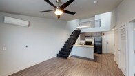This contemporary two-story space is designed with a modern aesthetic and an efficient, open layout. The main floor showcases a bright and airy living area with warm wood-look flooring, clean white walls, and a ceiling fan that adds both comfort and style. The open-concept design flows seamlessly into the kitchen, which features gray cabinetry, a light-toned countertop peninsula, and black appliances including a refrigerator and range. The peninsula serves as a natural divider between the living and kitchen areas, making the space feel connected yet purposefully defined. A striking staircase with dark treads and sleek metal railings leads to the upper level, drawing the eye upward and emphasizing the vertical scale of the space. The upper loft level is visible from below, highlighted by a horizontal metal railing that adds an industrial-modern touch to the overall design. The double-height ceiling in the main living area enhances the sense of openness, allowing light to travel freely throughout. The combination of wood-look flooring, neutral tones, and contemporary fixtures gives the space a clean, cohesive, and inviting character suited for modern living.