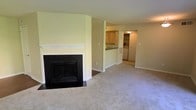 This single-floor residence offers a clean, functional layout designed for comfortable everyday living. The living room serves as the heart of the home, featuring plush wall-to-wall carpeting and a classic wood-mantled fireplace with a dark surround, adding a warm and inviting focal point to the space. Soft, neutral-toned walls throughout create a bright and airy atmosphere that complements the open feel of the floor plan. A pass-through opening connects the living area to the kitchen, providing an open-concept flow that enhances both functionality and visibility between spaces. The kitchen is outfitted with warm wood cabinetry, ample counter space, and a raised bar-height counter ideal for casual dining or meal prep. A dedicated laundry area with a full-size washer and dryer is conveniently situated just off the kitchen, adding practical in-unit utility. A built-in shelving unit nearby offers additional storage options. The overall design emphasizes ease of living with its straightforward, well-connected layout, neutral finishes, and essential amenities all contained within a single, accessible level.
