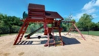 Located at 51 Northtown Dr in Jackson, Mississippi, this single-floor property includes a notable outdoor recreational area centered around a large, multi-feature wooden playground structure. The play structure is finished in a deep red tone and incorporates a variety of engaging elements, including a covered upper platform with a slatted wood roof, a tunnel slide, a traditional slide, multiple swings, climbing ladders, and tire accessories. The structure is set within a sandy ground surface that provides a natural safety buffer, and the entire play area is enclosed by a chain-link fence that defines the space and ensures a contained environment. Mature trees and dense greenery line the perimeter, offering natural shade and a lush backdrop that enhances the outdoor atmosphere. The setting benefits from open sky exposure, making it a well-lit and inviting outdoor space. The combination of varied play equipment and the surrounding natural landscape creates an active and functional recreational environment well-suited for organized outdoor use.