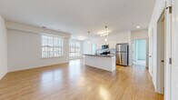 This single-floor residence is designed with an open-concept layout that seamlessly connects the living area and kitchen, creating a spacious and airy atmosphere. Warm-toned hardwood flooring extends throughout the main living spaces, reflecting abundant natural light that pours in through large, blinds-fitted windows overlooking the surrounding neighborhood.

The kitchen serves as a central focal point, anchored by a substantial island with dark granite countertops and a gold-finished faucet. White shaker-style cabinetry lines the walls, complemented by a classic subway tile backsplash and a full suite of stainless steel appliances, including a gas range, microwave, and refrigerator. Pendant lighting above the island and a track light fixture add both task and ambient illumination.

The bedroom features plush carpeting and a generously sized window, maintaining the home's bright, neutral palette. Two bathrooms are present — one with a walk-in shower and wood-look tile flooring, the other with a full shower-tub combination. An in-unit laundry closet houses a full-sized washer and dryer with overhead shelving, adding everyday convenience to this well-appointed residence.
