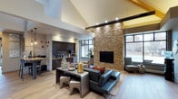 This single-floor space makes a striking first impression with its soaring vaulted ceilings accented by exposed dark wood beams, creating an open and airy atmosphere throughout. The open-concept layout connects the living and kitchen areas seamlessly, promoting a natural flow ideal for both everyday living and entertaining.

The living area is anchored by a dramatic floor-to-ceiling stacked stone fireplace wall, which serves as a bold architectural focal point. A flat-screen television is mounted within the stonework, blending functionality with design. Sleek leather seating and a studded industrial-style coffee table with matching ottomans add a contemporary edge to the space, while a textured area rug grounds the seating arrangement warmly.

Large windows on multiple walls invite generous natural light, offering views of the surrounding outdoor landscape and enhancing the bright, welcoming ambiance of the interior.

The kitchen, visible from the living area, features dark espresso cabinetry, modern appliances, and a tile backsplash, maintaining a cohesive aesthetic. Light hardwood flooring runs throughout, tying the spaces together with warmth and continuity.

Overall, this space balances rustic elements — exposed beams, natural stone — with modern finishes and furnishings, resulting in a refined yet comfortable interior.