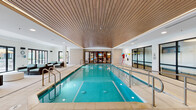 This indoor pool facility at Riverwalk in Plainsboro, NJ offers a well-appointed aquatic amenity space designed for both recreation and relaxation. The centerpiece is a generously sized lap pool with crystal-clear turquoise water, outfitted with stainless steel entry railings and underwater lighting that enhances the inviting atmosphere. Above the pool, a distinctive slatted wood ceiling adds warmth and architectural character, contrasting elegantly with the surrounding white walls and light-toned tile flooring.

The pool area is flanked by a spacious lounge zone furnished with wicker armchairs and light cushions, providing a comfortable retreat for residents. Large glass doors and windows along one side of the room allow natural light to filter in and offer views to an outdoor area, contributing to an open, airy ambiance throughout the space.

A striking blue mosaic tile accent wall serves as a bold focal point near the pool entry, complemented by potted tropical plants that introduce a touch of greenery. The facility includes a dedicated entry corridor with additional seating and direct exterior access, making it easily accessible. Safety signage and equipment are thoughtfully integrated without disrupting the refined aesthetic, resulting in a polished, resort-inspired environment suited to everyday use.