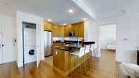 This single-floor space centers around a well-appointed, modern kitchen that balances functionality with contemporary style. The kitchen is outfitted with warm honey-toned wood cabinetry that extends to the ceiling, providing generous storage throughout. Dark granite countertops offer a striking contrast against the lighter cabinetry, running along the perimeter and across a central island that doubles as a breakfast bar. The island accommodates multiple white and chrome bar stools, making it an ideal spot for casual dining or entertaining.

Stainless steel appliances, including a full-size refrigerator and built-in microwave, complement the kitchen's modern aesthetic. Recessed lighting in the white ceiling ensures the space is well-lit, while light hardwood flooring flows throughout, contributing to a warm and cohesive look.

A discreet in-unit laundry area is tucked behind louvered doors just off the kitchen, adding everyday convenience without disrupting the overall design. An adjacent doorway hints at additional living areas beyond, suggesting a thoughtfully laid-out floor plan. The overall ambiance is clean, modern, and practical.