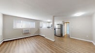 This single-floor unit offers a clean, functional layout with a bright and open feel throughout. The main living area is generously sized, featuring warm luxury vinyl plank flooring, neutral-toned walls, and a wall-mounted air conditioning unit alongside baseboard heating for year-round comfort. Natural light enters through multiple windows fitted with blinds.

The kitchen is positioned in a semi-open layout with a half-wall peninsula that connects it to the living space. It is equipped with stainless steel appliances — including a refrigerator, electric range, built-in microwave, and dishwasher — complemented by white upper cabinetry, grey lower shaker-style cabinets, light countertops, and a classic white subway tile backsplash. A window above the sink adds natural light to the workspace.

The bedroom is carpeted and comfortably sized, with a window providing natural light and a built-in closet with shelving for ample storage. Baseboard heating is also present in this room.

The bathroom is finished in a crisp, all-white palette, featuring a shower-tub combination with tiled surround, a vanity with under-sink storage, and luxury vinyl plank flooring. The overall aesthetic of the unit is clean and modern, presenting a well-maintained and move-in ready space.