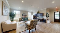 This single-floor combined kitchen and living room offers a well-organized, open layout that efficiently integrates two functional areas. The living area is anchored by neutral-toned walls, crown molding, and recessed lighting overhead, creating a clean and welcoming atmosphere. Wood-style flooring runs throughout the entire space, providing visual continuity. Large, multi-pane windows along multiple walls flood the room with natural light and offer views of the surrounding greenery outdoors. The living zone features a comfortable seating arrangement with patterned accent chairs, a cream sofa, and a woven area rug, complemented by a dedicated coffee station housed within a classic wooden armoire. The kitchen area is positioned along one wall and is equipped with dark espresso cabinetry, granite-style countertops, a mosaic tile backsplash, a stainless steel refrigerator, and a sink. A small dining table with metal chairs bridges the kitchen and living areas. A teal accent cabinet near the windows adds a pop of color to the overall warm, neutral palette. Together, these elements create a cohesive, inviting, and practical communal space.