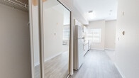 This single-floor studio unit offers a thoughtfully arranged layout that maximizes its compact footprint. The open-plan kitchen and living area form the heart of the space, where light hardwood-style flooring extends throughout the kitchen zone, transitioning to plush, neutral-toned carpet in the living area. The kitchen is equipped with white cabinetry featuring gold-tone trim, a white electric range, over-range microwave, and a standard sink set into a laminate countertop. A large window positioned above the countertop draws in generous natural light, brightening the entire space. A mirrored sliding closet door near the entry reflects additional light and contributes to a sense of openness.

The living area benefits from a notably vaulted ceiling with a ceiling fan, adding vertical dimension and improving airflow. Multiple windows fitted with horizontal blinds allow controllable natural light throughout the day. A wall-mounted unit provides climate control. The bathroom includes a wide vanity with an extended countertop, under-sink cabinetry, a large mirror, and a shower/tub combination. Neutral beige tones throughout maintain a clean, cohesive aesthetic across the entire unit.