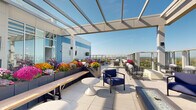 This expansive rooftop terrace offers a thoughtfully designed outdoor amenity space that blends contemporary architecture with a welcoming, open-air atmosphere. The terrace is anchored by a sleek pergola framework that provides partial shade while preserving unobstructed views of the surrounding treetops, skyline, and a distant waterway visible on the horizon.

The outdoor space is organized into multiple distinct seating zones, each furnished with modern lounge chairs, sectional sofas, bistro-style tables, and accent side tables. A built-in linear gas fireplace is mounted into the exterior building façade, accompanied by a wall-mounted display screen, creating an inviting focal point for gatherings in cooler weather. Floor-to-ceiling glass walls connect the terrace to an interior lounge area, allowing natural light to flow freely between the indoor and outdoor spaces.

Vibrant planter boxes filled with colorful seasonal flowers—including chrysanthemums in warm yellows, oranges, and purples—add lively bursts of color throughout the terrace. Large-format light-toned pavers cover the entire deck surface, contributing to the clean, contemporary aesthetic. Glass panel railings line the perimeter, ensuring uninterrupted panoramic views from every seating area. The overall ambiance is bright, airy, and elevated, offering a sophisticated retreat above the cityscape.