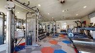 This fitness center is a fully equipped workout facility designed to accommodate a wide range of exercise routines. The space is organized into distinct functional zones, seamlessly flowing from one area to the next across a single floor. A striking hexagonal-patterned floor in red, blue, and grey tones adds a bold, energetic aesthetic throughout the entire space.

The strength training area is comprehensively outfitted with a full dumbbell rack, a multi-function cable machine, a Smith machine, and dedicated free weight stations. Floor-to-ceiling mirrors line portions of the walls, enhancing the sense of space and allowing users to monitor their form. Kettlebells and weight plates are neatly organized and readily accessible.

The cardio zone features a row of treadmills, elliptical machines, and a stationary spin bike, positioned near large windows that allow natural light to fill the space. A wall-mounted television is also present for entertainment during workouts.

One wall is adorned with a vibrant, large-scale graphic mural in red and teal, branded with the "Club Sweat" name, adding a dynamic visual focal point to the space. Glass doors lead directly to an outdoor area, providing convenient access and natural ventilation. Recessed lighting throughout ensures the space remains well-lit at all times. An adjacent anteroom provides additional utility space.