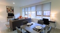 This single-floor urban residence is thoughtfully designed to maximize both comfort and functionality. The open-plan layout seamlessly connects the living and dining areas, creating a cohesive and inviting atmosphere throughout. Large windows fitted with roller shades line the living and dining space, flooding the interior with natural light while framing views of the surrounding cityscape. The dining area features a contemporary wood-toned table set for four, complete with place settings and glassware, making it well-suited for both everyday meals and entertaining guests. Adjacent to the dining area, the living room is furnished with a plush gray sofa, a vibrant orange accent chair, and decorative throw pillows, adding warmth and personality to the neutral, light-toned walls and flooring. A wall-mounted television and a classic table lamp contribute to the room's balanced blend of modern and traditional elements. The overall ambiance is clean, bright, and urban, reflecting a space that is both practical and stylish. The open flow between rooms enhances the sense of spaciousness, while the carefully chosen furnishings and natural light create a welcoming environment suited to a variety of lifestyles.