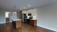 This single-floor space showcases a thoughtfully designed open-concept layout that emphasizes both style and functionality. The kitchen serves as a central highlight, featuring a striking two-tone cabinet design that pairs warm wood lower cabinets with crisp white upper cabinetry, creating a contemporary yet inviting aesthetic. A sleek kitchen island with a white countertop and an integrated sink provides additional workspace and a natural gathering point, accented by elegant pendant lighting that adds a warm, decorative touch overhead. The kitchen is equipped with stainless steel appliances, including a french door refrigerator, a microwave, and a gas range, all set against a clean subway tile backsplash that ties the design together seamlessly. Recessed lighting throughout ensures the space is well-lit and open. The rich, dark hardwood flooring runs continuously across the space, grounding the design and adding a sense of warmth and sophistication. Neutral gray walls provide a versatile backdrop that complements the bold cabinetry and flooring choices. The overall ambiance is modern and polished, with a clean, uncluttered feel that makes the space feel expansive and welcoming.