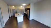 This single-floor space showcases a thoughtfully designed open-concept layout, where the kitchen and living area flow seamlessly together. The kitchen is a standout feature, equipped with dark espresso-toned cabinetry that provides generous upper and lower storage. Stainless steel appliances, including a refrigerator, electric range, and built-in microwave, complement the clean white countertops. A tile backsplash adds a subtle textural detail behind the cooking area. A central island with an integrated sink serves as both a practical workspace and a natural dividing point between the kitchen and the open living space, enhancing the room's functionality.

The living area is spacious and open, finished with rich, dark hardwood-style flooring that extends throughout, adding warmth and visual continuity. A ceiling fan is installed overhead, contributing to the room's comfort. Flush-mount lighting fixtures illuminate both the kitchen and adjoining spaces effectively. Paneled gray interior doors are featured throughout, adding a clean, finished look. The neutral wall tones and open layout create a bright, airy ambiance that makes the space feel welcoming and versatile.