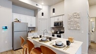 This contemporary single-floor residence offers a thoughtfully designed layout that blends modern style with everyday functionality. The open-concept kitchen is a standout feature, showcasing white shaker cabinetry, grey subway tile backsplash, stainless steel appliances including a range, microwave, and refrigerator, and a sleek white quartz peninsula with an undermount sink and seating for casual dining. Cylindrical pendant lights add a refined touch overhead.

The living area is bright and welcoming, with light hardwood-style flooring, elevated ceilings, and a glass-paneled exterior door that fills the space with natural light and provides direct outdoor access. The bedroom offers plush carpeting, soft neutral tones, ample natural light through large windows with sheer curtains, and thoughtful wall accents including decorative round mirrors.

Two bathrooms serve the residence, each finished with white cabinetry, quartz countertops, and tub-shower combinations. One features wood-look tile flooring and a double vanity, while the other offers a single vanity with well-lit mirror. Throughout, the design maintains a cohesive, clean aesthetic with neutral palettes, modern fixtures, and carefully considered details that create a comfortable and stylish living environment.