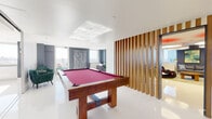 This thoughtfully designed single-floor space combines contemporary aesthetics with functional versatility across several distinct areas. The centerpiece of the space is a generously sized billiards room, anchored by a solid wood pool table with a rich burgundy felt surface. A striking vertical wood slat partition elegantly defines this area while maintaining an open, airy connection to adjacent rooms. Glossy white flooring runs throughout, reflecting abundant natural light that pours in from large windows offering sweeping panoramic city views.

The open-concept kitchen is sleek and modern, featuring dark cabinetry, a contrasting dark-tiled backsplash, a central island with bar seating, built-in appliances, and a spacious dining table accommodating a large group. Track lighting overhead ensures the area is well-lit for both cooking and gathering.

A casual lounge area offers a comfortable sectional sofa set against blue-grey accent walls, with curated artwork adding a creative touch. A separate, more intimate lounge features bold navy blue walls, sculptural statement furniture, oversized pendant ceiling fixtures with red accents, and floor-to-ceiling windows that frame impressive city skyline views. Together, these spaces create a dynamic, design-forward environment ideal for both social gatherings and relaxed lounging.