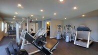 This fitness center is a thoughtfully designed workout facility that provides a comprehensive exercise environment for users of all fitness levels. The space is outfitted with professional-grade Precor USA equipment, including a leg press machine, multiple elliptical trainers, and a cable/pulley strength system, ensuring a full range of strength and cardiovascular training options. The room features durable dark rubber flooring, ideal for heavy equipment use and providing both safety and easy maintenance. Neutral gray walls create a clean, focused atmosphere, while recessed ceiling lights illuminate the space evenly throughout. An adjacent room, visible through an open doorway, appears to serve as a changing or restroom facility, adding convenience for users. The layout is organized to maximize the available floor space, allowing for comfortable movement between machines. The overall ambiance is practical and functional, reflecting a well-maintained facility designed to support a consistent and effective fitness routine.
