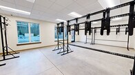Located at 133 S East St in Amherst, Massachusetts, this single-floor fitness studio offers a well-designed, open training environment purpose-built for functional exercise. The space features a polished concrete floor that provides a durable, easy-to-maintain surface suitable for a wide range of workout activities. Multiple freestanding and wall-mounted black steel functional training rigs dominate the layout, offering attachment points for resistance bands, suspension trainers, and other fitness accessories, making the studio highly versatile for strength and conditioning workouts. The drop ceiling is fitted with recessed fluorescent panel lighting, ensuring consistent, even illumination throughout. A wall-mounted mini-split HVAC unit provides climate control, keeping the environment comfortable year-round. Large windows on two walls flood the studio with natural light while offering views of the surrounding streetscape, creating an open and energizing atmosphere. A glass-paneled entry door with an illuminated exit sign ensures safe and easy access. The neutral, cream-colored walls contribute to a clean, professional aesthetic. The overall layout is efficient and functional, maximizing usable floor space for training while maintaining an uncluttered, motivating environment ideal for personal training, small group fitness classes, or specialty programming.