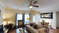 This single-story space combines comfort and functionality in a thoughtfully designed open-concept layout. The living area serves as the centerpiece, anchored by a plush, neutral-toned sofa adorned with decorative throw pillows and complemented by a classic brown leather accent chair. A ceiling fan with light fixture overhead provides both ambient lighting and air circulation. Crown molding adds an elegant architectural detail along the upper walls, enhancing the room's refined character.

Natural light flows in through multiple windows, including a prominent bay window dressed with soft blue curtains and slatted blinds, creating a bright and inviting atmosphere. A glass-top coffee table sits atop a patterned area rug, adding a reflective elegance to the space. Track lighting along the ceiling offers additional illumination options.

The living area transitions seamlessly into a dining space, where a wooden table surrounded by upholstered chairs is visible. A framed mirror on the wall enhances the sense of depth, while a decorative floral arrangement and tabletop accents add warmth. Rich hardwood flooring runs throughout, tying the entire space together with a classic, polished aesthetic.