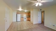 This single-floor residence offers a practical and well-organized open-concept layout that seamlessly connects the living area and kitchen into one cohesive space. The kitchen is equipped with light wood cabinetry providing ample storage, a white gas range, a full-size refrigerator, and a built-in range hood. The countertops and lower cabinetry wrap around in an L-shaped configuration, making efficient use of the available space. Large neutral-toned ceramic floor tiles extend throughout the kitchen and main living area, lending a clean and cohesive look that is easy to maintain. The living area transitions into a carpeted zone, adding a touch of warmth and comfort underfoot. A ceiling fan with an integrated light fixture is centrally positioned overhead, ensuring good air circulation and ambient lighting across the open floor plan. Additional overhead fluorescent lighting in the kitchen area provides bright, functional illumination for cooking and meal prep. The space includes multiple interior doors leading to additional rooms, as well as a visible HVAC vent, contributing to a comfortable interior climate. The overall ambiance is bright, neutral, and move-in ready, offering a functional layout suitable for everyday living.