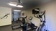This single-floor fitness center is designed to provide a functional and comfortable workout environment. The space is equipped with a thoughtfully curated selection of True-brand cardio machines, including a recumbent bike, an upright stationary bike, and an elliptical trainer, catering to a range of cardiovascular exercise preferences. A multi-function cable weight machine paired with a workout bench occupies the far end of the room, offering strength training options for users. The textured ceiling is fitted with a ceiling fan and overhead lighting, ensuring adequate airflow and illumination throughout workouts. A wall-mounted flat-screen television is positioned for easy viewing from the cardio equipment, adding an entertainment element to exercise sessions. The flooring features dark, durable gym-grade material suited for high-traffic use. The walls are painted in a neutral blue-gray tone, contributing to a calm and focused atmosphere. Motivational fitness posters accent the walls, rounding out the space with an encouraging visual touch. Overall, the fitness center is a practical, no-frills facility well-suited for everyday exercise routines.
