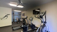 This single-floor fitness center is designed to provide a functional and comfortable workout environment. The space is equipped with a thoughtfully curated selection of True-brand cardio machines, including a recumbent bike, an upright stationary bike, and an elliptical trainer, catering to a range of cardiovascular exercise preferences. A multi-function cable weight machine paired with a workout bench occupies the far end of the room, offering strength training options for users. The textured ceiling is fitted with a ceiling fan and overhead lighting, ensuring adequate airflow and illumination throughout workouts. A wall-mounted flat-screen television is positioned for easy viewing from the cardio equipment, adding an entertainment element to exercise sessions. The flooring features dark, durable gym-grade material suited for high-traffic use. The walls are painted in a neutral blue-gray tone, contributing to a calm and focused atmosphere. Motivational fitness posters accent the walls, rounding out the space with an encouraging visual touch. Overall, the fitness center is a practical, no-frills facility well-suited for everyday exercise routines.