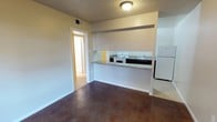 This single-floor unit offers a straightforward, functional layout designed for efficient everyday living. The main living area features stained concrete floors that lend an industrial warmth to the space, complemented by neutral-toned walls and a textured ceiling with flush lighting overhead. A defining feature of the layout is the open kitchen pass-through, framed by a half-wall with a granite-style countertop that connects the kitchen to the living area. The kitchen is equipped with essential appliances including a white electric range, a sink, and a full-size refrigerator, all set against dark lower cabinetry that provides a subtle contrast to the lighter walls. A HVAC vent is visible near the ceiling, indicating central climate control throughout. A doorway off the main living space leads further into the unit, suggesting additional rooms beyond — likely a bedroom and bathroom. The overall ambiance is clean and neutral, offering a blank canvas that is easy to personalize. The space is modest in scale but well-organized, making practical use of every square foot available.