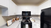 This single-floor residence offers a thoughtfully designed living space centered around a clean and functional kitchen. The kitchen showcases crisp white shaker-style cabinetry with sleek brushed nickel hardware, providing ample storage and a bright, timeless aesthetic. Light-toned countertops offer generous prep space, while the decorative mosaic tile backsplash in neutral gray tones adds a modern touch and visual interest to the cooking area. Black appliances, including an electric range and an over-the-range microwave, create a bold contrast against the white cabinetry and lighter surfaces. The kitchen layout is efficiently arranged, making the most of the available space for both cooking and organization. A textured ceiling adds a subtle architectural detail throughout the home. The overall ambiance of the space is clean, practical, and inviting, with a neutral color palette that lends itself well to a variety of décor styles. The thoughtful combination of classic white finishes and contemporary accents gives this home a balanced and welcoming character suitable for everyday living.