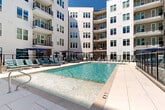 Located at 3310 S Tryon St in Charlotte, NC, this outdoor amenity space offers a resort-style pool courtyard that spans a generous footprint across a single level. The centerpiece is a large, rectangular swimming pool with crystal-clear blue water, framed by wide, light-toned tile decking that provides ample room for relaxation and movement. The sundeck is well-appointed with numerous cushioned lounge chairs in teal upholstery, accompanied by bold navy blue market umbrellas that provide shade throughout the day. A decorative water feature with smooth river rocks and sleek glass panel fencing adds a refined architectural detail along one edge of the pool area. The space is enclosed within a stylish courtyard formed by a modern multi-story building clad in crisp white siding, with large windows and private balconies overlooking the amenity below. A black metal perimeter fence defines the pool zone, ensuring both safety and visual structure. The open-sky setting allows abundant natural light to fill the courtyard, creating a bright and inviting atmosphere. Accessible entry points and clean sightlines throughout the space make it both functional and aesthetically cohesive, ideal for outdoor leisure and community gathering.