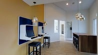 This contemporary single-floor space is thoughtfully designed to balance function and style. The business center area is a standout feature, showcasing sleek, wall-mounted workstations with integrated monitors in a striking navy and white finish, each paired with upholstered black leather bar stools. Bold mustard-yellow accent walls contrast with crisp white surfaces, lending the space a vibrant yet professional energy. Gleaming gold globe pendant lights hang from the soaring vaulted ceiling, adding a touch of sophistication and drawing the eye upward to highlight the impressive vertical scale of the room. Light wood-style flooring runs throughout, providing a warm, cohesive foundation that complements the modern aesthetic. Recessed lighting supplements the pendant fixtures, ensuring the space is well-illuminated throughout. Adjacent to the workstation area, a glimpse of an additional room is visible through a glass-paneled door, hinting at further amenities beyond. A high-top bar or counter area is also visible toward the right, offering additional seating and flexible use. The overall ambiance is modern, energetic, and purposefully designed to support productivity and comfort.