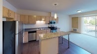 This single-floor unit offers a thoughtfully designed layout that balances functionality and comfort. The kitchen serves as the heart of the space, showcasing light maple cabinetry, dark countertops, and a full suite of stainless steel appliances including a refrigerator, electric range, built-in microwave, and dishwasher. A central granite-topped island with sleek metal legs provides additional prep space and doubles as a casual dining area, illuminated by a stylish pendant light overhead. Rich hardwood-style flooring runs throughout the kitchen and main living areas, adding warmth and visual continuity.

The bedroom is a bright and comfortable retreat, featuring plush wall-to-wall carpeting, a ceiling fan with integrated light fixture, and two windows that frame views of the surrounding urban cityscape, allowing natural light to fill the room.

The bathroom is clean and practical, featuring wood-style flooring that complements the rest of the unit, a combined bathtub and shower with chrome fixtures, a toilet, and a vanity with ample counter space. Light-toned cabinetry and neutral wall colors create a calm, cohesive aesthetic throughout the space.

Overall, the unit presents a modern, move-in-ready living environment with a clean design palette and city-facing outlook.