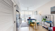 This thoughtfully designed studio unit combines functional living with contemporary style across a single, open-concept floor. Light wood flooring and crisp white walls run throughout, creating a clean and airy aesthetic that is further enhanced by an abundance of natural light streaming through large, floor-to-ceiling windows overlooking a surrounding urban skyline.

The main living area seamlessly integrates sleeping, dining, and lounging zones. A full-size bed anchors the sleeping area, positioned directly in front of the expansive windows to maximize the view. A compact dining nook with a round white table and eclectic seating sits nearby, while a ceiling fan overhead keeps the space comfortable year-round. Recessed lighting adds a polished finish to the ceiling.

The kitchen is efficiently tucked along one wall and features a full suite of stainless steel appliances, including a refrigerator, range, microwave, and dishwasher. Light wood upper cabinetry pairs with grey lower cabinets and a tiled backsplash for a modern, two-tone look.

A built-in closet with an organized shelving system provides practical storage. The bathroom is finished with large-format marble-look floor and wall tiles, a floating grey vanity with a white countertop, and matte black fixtures, lending a sleek, upscale feel to the space.