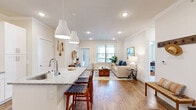 This thoughtfully designed single-floor residence offers a seamless open-concept layout that connects the kitchen and living room, creating a bright and cohesive main living area. The kitchen is a standout feature, showcasing white shaker-style cabinetry, granite countertops, stainless steel appliances, and a peninsula with bar seating illuminated by sleek pendant lighting — ideal for both meal preparation and casual dining. Warm hardwood flooring flows throughout the main living areas, complementing the neutral, light-toned walls and crown molding details that lend an air of refinement. The living room is generously proportioned, bathed in natural light from large windows, and features recessed lighting overhead. The primary bedroom offers a comfortable, carpeted retreat with a ceiling fan and ample space. The full bathroom is finished with a granite-topped double vanity, white cabinetry, and wood-look flooring. A dedicated office provides a quiet, flexible workspace, while two closets offer practical storage solutions. A hallway area with natural light and sheer window treatments adds an airy transition between spaces. Throughout, the design maintains a clean, modern aesthetic with warm accents, making this residence both functional and inviting.