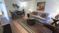 This two-story space offers a welcoming open-concept layout where the living and dining areas flow seamlessly together. The main living area features warm hardwood flooring that extends throughout, creating a cohesive and inviting atmosphere. A neutral color palette on the walls provides a versatile backdrop, complemented by colorful accents including decorative throw pillows in deep reds and earthy tones.

The living area is anchored by a generously sized gray sofa adorned with patterned and solid accent pillows, paired with a rustic wooden coffee table topped with a decorative bowl. A patterned area rug with bordered detailing defines the seating space, adding texture and warmth underfoot. A vibrant floral canvas artwork serves as a focal point on the main wall, adding visual interest to the room.

Adjacent to the living space, the dining area accommodates a dark wood table surrounded by a mix of wooden and wicker-style chairs, ideal for everyday dining. Decorative plates arranged artistically on the wall add character to this corner. Ambient lighting from a floor lamp and table lamp enhances the overall warm, comfortable ambiance of the space.