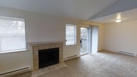 This single-floor space centers around a comfortable living area defined by a tile-surround fireplace with a clean white mantel, serving as a focal point along the main wall. The flooring transitions from ceramic tile near the fireplace to wall-to-wall carpeting throughout the remainder of the living area, providing a warm and practical surface underfoot. Natural light enters the room through a window fitted with horizontal blinds, while a sliding glass door with sheer curtains opens to an outdoor balcony, offering views of surrounding trees and extending the sense of space beyond the interior. A baseboard heating unit runs beneath the window, ensuring efficient climate control throughout the room. The neutral color palette—encompassing light beige walls and cream-toned ceilings—creates a bright, understated ambiance that is both versatile and welcoming. The open layout of the living area allows for flexible furniture arrangement, and the combination of the fireplace, balcony access, and natural light contributes to an overall atmosphere that is both functional and comfortable for everyday living.