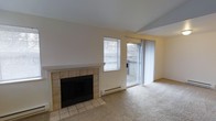 This single-floor space centers around a comfortable living area defined by a tile-surround fireplace with a clean white mantel, serving as a focal point along the main wall. The flooring transitions from ceramic tile near the fireplace to wall-to-wall carpeting throughout the remainder of the living area, providing a warm and practical surface underfoot. Natural light enters the room through a window fitted with horizontal blinds, while a sliding glass door with sheer curtains opens to an outdoor balcony, offering views of surrounding trees and extending the sense of space beyond the interior. A baseboard heating unit runs beneath the window, ensuring efficient climate control throughout the room. The neutral color palette—encompassing light beige walls and cream-toned ceilings—creates a bright, understated ambiance that is both versatile and welcoming. The open layout of the living area allows for flexible furniture arrangement, and the combination of the fireplace, balcony access, and natural light contributes to an overall atmosphere that is both functional and comfortable for everyday living.